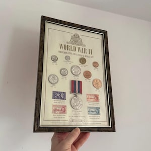 Puede incluir: Conjunto enmarcado de monedas, sellos y medallas conmemorativas de la Segunda Guerra Mundial. La exhibición incluye varias monedas, sellos y una medalla con cintas rojas, blancas y azules. El marco es marrón oscuro.