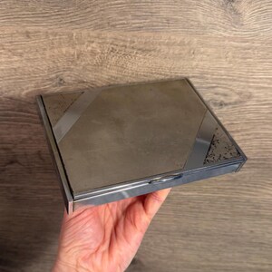 May include: A rectangular silver-toned metal compact case featuring a smooth, matte-finish center panel flanked by two polished, reflective diagonal sections. The edges are finished in a high-shine chrome, and a small latch is visible on the front.