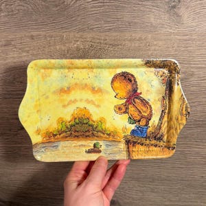 May include: A rectangular decorative tray featuring a cartoon bear in a red scarf and blue jeans, gazing at a duck on a pond. The tray has a yellow background with a landscape scene.