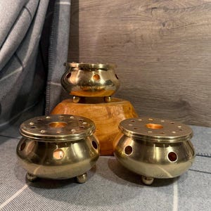 Puede incluir: Un conjunto de tres quemadores de incienso de latón. Dos quemadores tienen una tapa redonda perforada y tres patas pequeñas. El tercer quemador, más pequeño, con un diseño similar, se encuentra sobre una base de madera. Los quemadores tienen un tono dorado cálido.