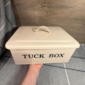 Könnte beinhalten: Cremefarbene rechteckige Metall-Aufbewahrungsbox mit passendem Deckel und gebogenem Griff. Die Worte "TUCK BOX" sind in Schwarz auf die Vorderseite der Box gedruckt. Die Box wird von einer Hand gehalten.