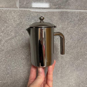 May include: A stainless steel French press coffee maker with a handle and a lid. The coffee maker is cylindrical with a spout for pouring. The lid has a small decorative knob on top. The coffee maker is held in a hand.