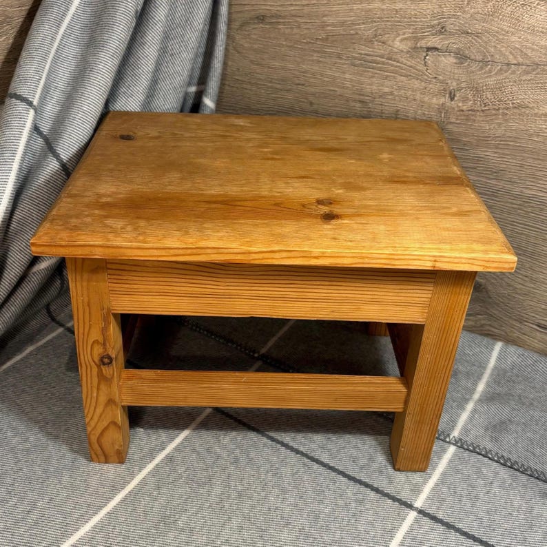 May include: A small, square wooden step stool with a natural finish. The stool has four sturdy legs and a simple, functional design. The wood grain is visible, adding to its rustic charm. The stool measures approximately 12 inches in height.