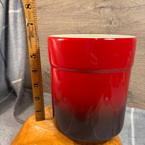May include: A red and black ceramic utensil holder. The holder is cylindrical and has a gradient color scheme, transitioning from red to black.