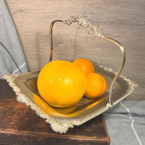 Puede incluir: Una cesta de servir cuadrada de color plateado con un asa decorativa y bordes ornamentados. La cesta contiene tres naranjas. El asa tiene un adorno decorativo en la parte superior. La cesta está sobre una superficie marrón.
