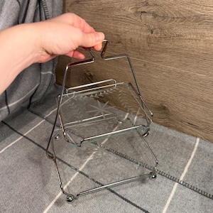 May include: A two-tiered serving tray with a silver metal frame and etched glass shelves. The glass shelves feature a star and leaf design. The tray is held by a handle at the top.