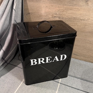May include: A glossy black metal bread bin with a rectangular shape and rounded corners. The word BREAD is printed in white capital letters on the front. It features a removable lid with a curved black handle centred on the top surface.