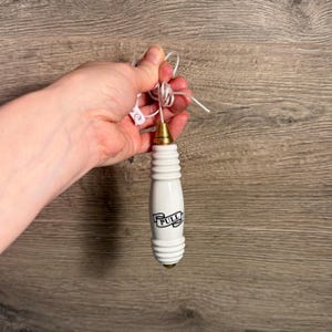 Op de afbeelding: Een witte keramische trekhendel met een goudkleurige basis en een wit koord. Op de hendel staat het woord "PULL" in het zwart gedrukt. De hendel wordt tegen een houten achtergrond gehouden.