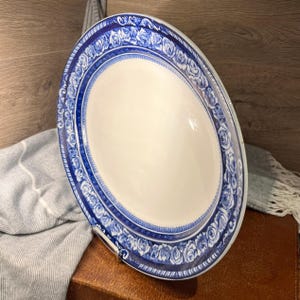 May include: An oval serving platter with a white centre and a blue floral border. The blue border features a repeating rose pattern. The platter is made of ceramic and is displayed on a brown surface.