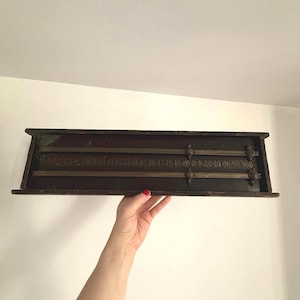 Puede incluir: Un antiguo dispositivo de medición de madera y latón con números y líneas. El dispositivo tiene un marco de madera marrón oscuro y barras de medición de latón. Los números están grabados en el latón. El dispositivo está en manos de una persona.