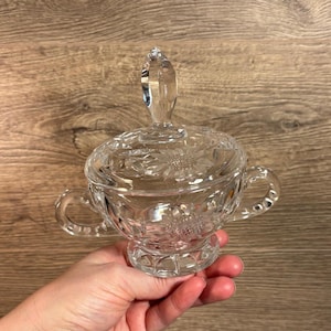 May include: A clear glass sugar bowl with a lid and two handles. The bowl is decorated with a floral pattern.