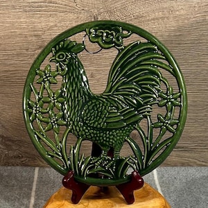 以下が含まれることがあります： 花と葉がついた雄鶏のデザインが施された緑色のセラミック製鍋敷き。鍋敷きはカットアウトデザインで、吊り下げ用のループが付いています。
