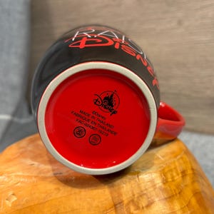 Peut inclure: Mug Disney en c&eacute;ramique noire avec int&eacute;rieur et anse rouges. Le mug porte le logo Disney et du texte au fond. L'ext&eacute;rieur pr&eacute;sente un texte rouge qui dit "RAISE DISNEY". Fabriqu&eacute; en Tha&iuml;lande.