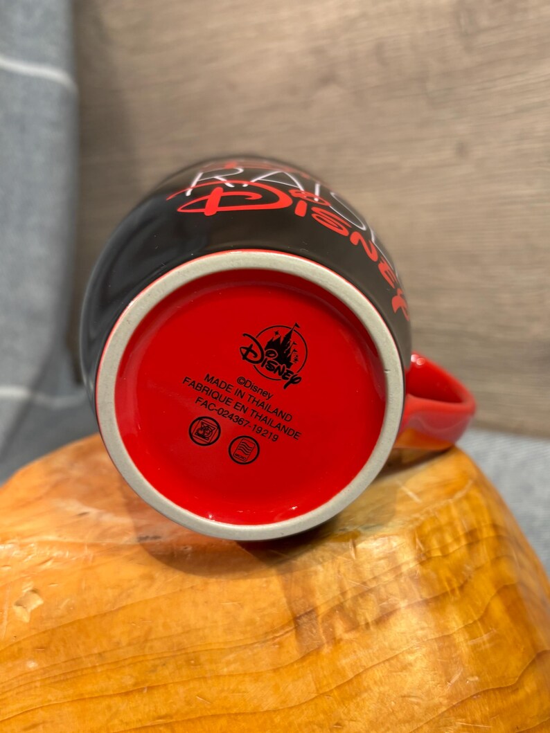 Peut inclure: Mug Disney en c&eacute;ramique noire avec int&eacute;rieur et anse rouges. Le mug porte le logo Disney et du texte au fond. L'ext&eacute;rieur pr&eacute;sente un texte rouge qui dit "RAISE DISNEY". Fabriqu&eacute; en Tha&iuml;lande.