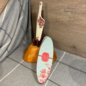 May include: A white cake server with a floral design, displayed on a wooden stand. The server features red and pink flowers. A light blue cake box with the text "Rosie's cake server" and a floral pattern is also present.