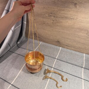 May include: A copper-colored incense burner with a decorative floral pattern, suspended by a gold chain. Two matching gold-colored handles are next to the burner. The burner is held by a hand against a gray and white plaid background.