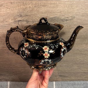 May include: Black ceramic teapot with a floral design. The teapot has a gold trim and handle, and a matching lid. The floral design features small white, orange, and blue flowers. The teapot is held by a hand.
