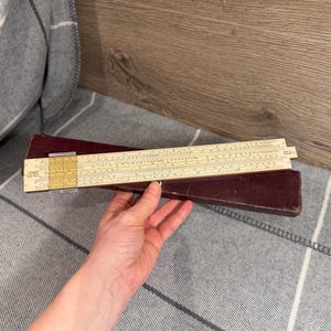 May include: A vintage slide rule with a cream-colored scale and a dark burgundy case. The slide rule is made of a light-colored material with black and red markings. The case is rectangular and appears to be made of leather or a similar material.
