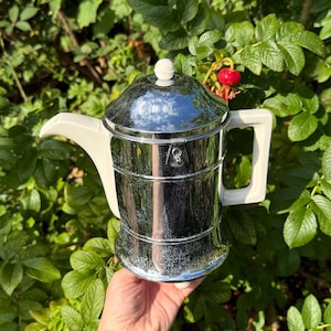 Puede incluir: Una cafetera cromada vintage con asa y boquilla blancas. La cafetera tiene una superficie brillante y reflectante y un pomo blanco en la tapa. La cafetera se sostiene frente a un fondo de hojas verdes.