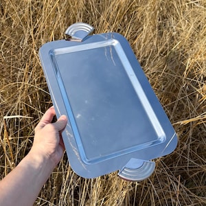 May include: A rectangular silver-toned metal serving tray with a polished, reflective surface. The tray features two semi-circular handles on opposite ends, each designed with a series of concentric, raised ridges for a textured grip.
