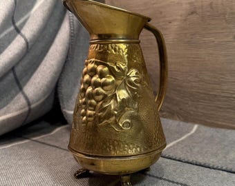 May include: A vintage brass pitcher with a textured surface and a grape vine design. The pitcher has a handle and is supported by three small claw feet. The spout is angled for easy pouring. The pitcher is likely used for serving beverages.