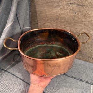 May include: An oval copper cooking pot with brass handles. The interior of the pot is tarnished with green and red colours. The exterior is a shiny copper colour. The pot is on a grey and white striped blanket.