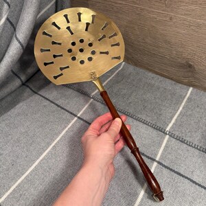 Puede incluir: Un espumadera de latón con un mango de madera oscura. La espumadera tiene una cabeza circular con formas decorativas recortadas y pequeños agujeros. El mango es largo y delgado, con un acabado pulido. Este utensilio de cocina está diseñado para colar alimentos.