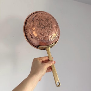 May include: A copper pan with a brass handle. The pan's interior has an embossed design, and the handle has a hole for hanging. The pan is a warm copper colour, and the handle is a shiny gold colour.