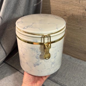 Könnte beinhalten: Ein weißer Keramikbehälter mit marmorähnlichem Muster in Blau und goldenen Akzenten. Der Behälter hat einen goldfarbenen Rand und einen goldenen Verschluss. Der Deckel ist rund und passt gut. Der Behälter dient wahrscheinlich zur Aufbewahrung von Trockenwaren.