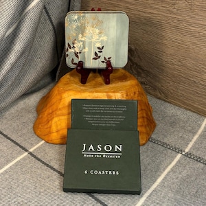 May include: A set of six coasters in a green box with the brand name "JASON" and the text "Make the Occasion". The coasters have a light blue background with gold floral designs and are displayed on a wooden stand.