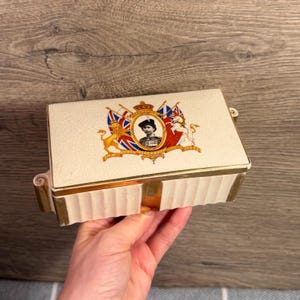 May include: A rectangular cream-colored ceramic box with a crackle glaze finish and gold-tone metal trim. The lid features a central oval portrait of Queen Elizabeth II, flanked by a lion and unicorn, with Union Jack flags and the text H.M. QUEEN ELIZABETH II.