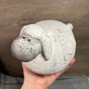 Könnte beinhalten: Eine weiße Keramik-Schaffigur mit einer strukturierten Oberfläche und einem grauen Gesicht. Das Schaf hält seinen Kopf gesenkt und hat zwei schwarze Augen.
