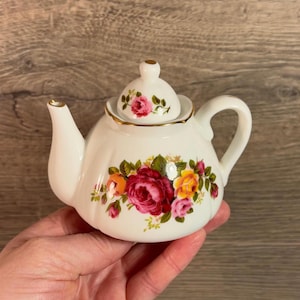 May include: A white porcelain teapot with a floral design featuring pink, yellow, and red roses. The teapot has a lid with a gold trim and a gold band around the top.