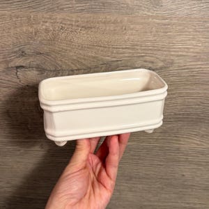 May include: A rectangular off-white ceramic container with a smooth, glossy finish. The vessel features a raised horizontal band detail along the exterior walls and is supported by four small, rounded feet at the base, creating a basin-like form.