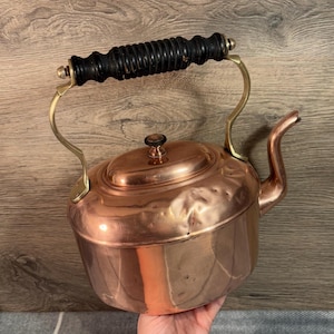 Può includere: Un bollitore in rame vintage con un manico in legno e dettagli in ottone. Il bollitore ha un corpo arrotondato e un beccuccio laterale. Il bollitore è leggermente ammaccato e mostra segni di età.