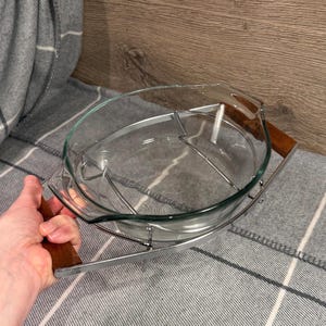 Peut inclure: Un plat à four ovale en verre transparent, avec un cadre métallique et des poignées en bois. Le cadre est argenté et les poignées sont brun foncé. Le plat est conçu pour être placé dans le cadre.