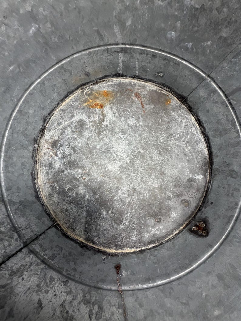 Op de afbeelding: De binnenkant van een ronde container van gegalvaniseerd metaal met een textuurrijk, grijs gevlekt oppervlak met zichtbare tekenen van oxidatie, waaronder kleine oranje roestvlekken en donkere plekken langs de ronde bodem.