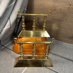 Pode incluir: Um conjunto de dois suportes de fogo de lareira de latão com um design decorativo. Os suportes de fogo estão empilhados um em cima do outro.