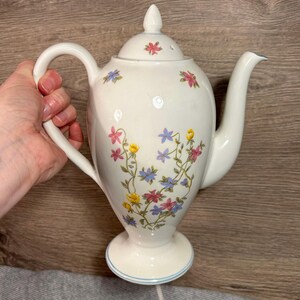 May include: White ceramic teapot with a floral design featuring pink, blue, and yellow flowers. The teapot has a lid with a small hole in the top and a handle on the side.
