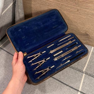 May include: An open, vintage drafting set in a brown case with a blue velvet interior. The set includes several silver-colored compasses, dividers, and other drawing tools. The case is held open by a hand.