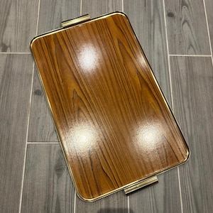 Puede incluir: Una bandeja rectangular de madera con ribete y asas de metal dorado. La bandeja tiene un patrón de veta de madera y un acabado brillante.