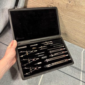 以下が含まれることがあります： 黒い革製のケースにベルベットの裏地が施され、銀色のコンパスがセットされています。コンパスは列に並べられており、さまざまなサイズとスタイルがあります。ケースは開いており、コンパスが見えます。