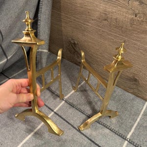 Peut inclure: Deux chenets en laiton. Chaque chenet a un dessus décoratif, un cadre rectangulaire et une base incurvée. Les chenets sont présentés sur une couverture à carreaux grise et blanche et une surface en bois.