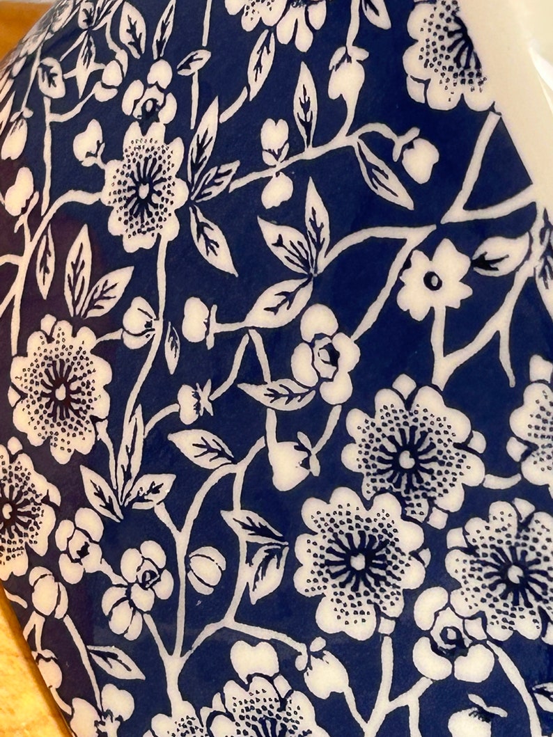 May include: A close-up of a blue and white ceramic mug with a floral pattern. The mug features a repeating design of white flowers and leaves on a dark blue background.