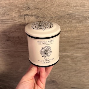 May include: A cylindrical cream-colored ceramic jar with a matching lid, featuring black text and circular logos. The front displays 'CROPWELL BISHOP CREAMERY', 'Potted BLUE STILTON with PORT', and a circular emblem with a tree and text.
