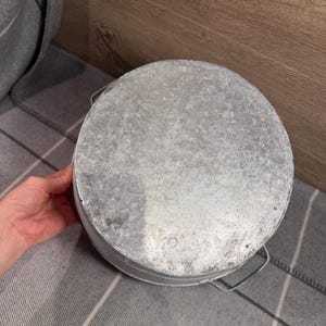 Op de afbeelding: Een ronde emmer van gegalvaniseerd metaal met een gestructureerde, gevlekte zilvergrijze afwerking. De container heeft een plat, cirkelvormig bovenoppervlak en dunne metalen draadhandvatten aan de zijkanten met een functioneel ontwerp.