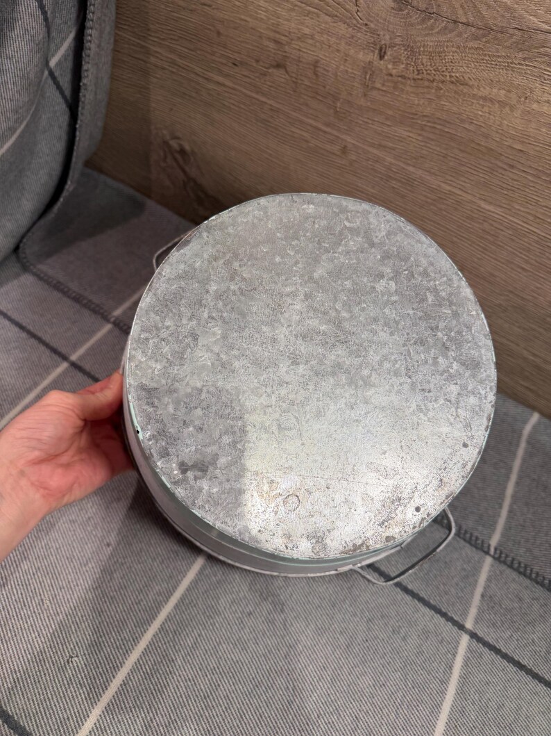 Op de afbeelding: Een ronde emmer van gegalvaniseerd metaal met een gestructureerde, gevlekte zilvergrijze afwerking. De container heeft een plat, cirkelvormig bovenoppervlak en dunne metalen draadhandvatten aan de zijkanten met een functioneel ontwerp.