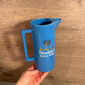 Könnte beinhalten: Ein hellblauer Keramikkrug mit einem quadratischen Griff und einem Ausguss. Die Vorderseite zeigt ein goldenes Wappenlogo über dem Text Embassy Number 1 Extra Mild, der in weißer und goldener Schrift auf dem glatten, zylindrischen Körper steht.