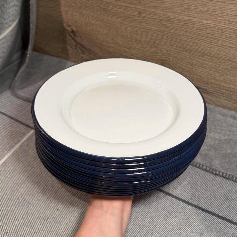 Pode incluir: Uma pilha de pratos redondos com uma superf&iacute;cie central branca lisa e brilhante e uma borda azul-marinho profunda. Os pratos t&ecirc;m um design circular limpo com um contraste de cores distinto entre o interior branco e as bordas externas escuras.