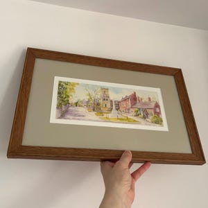 Puede incluir: Una pintura de acuarela enmarcada de una escena de pueblo. La obra de arte representa una iglesia con una torre, edificios y una calle. El marco es de madera marrón y el passe-partout es de color beige claro.